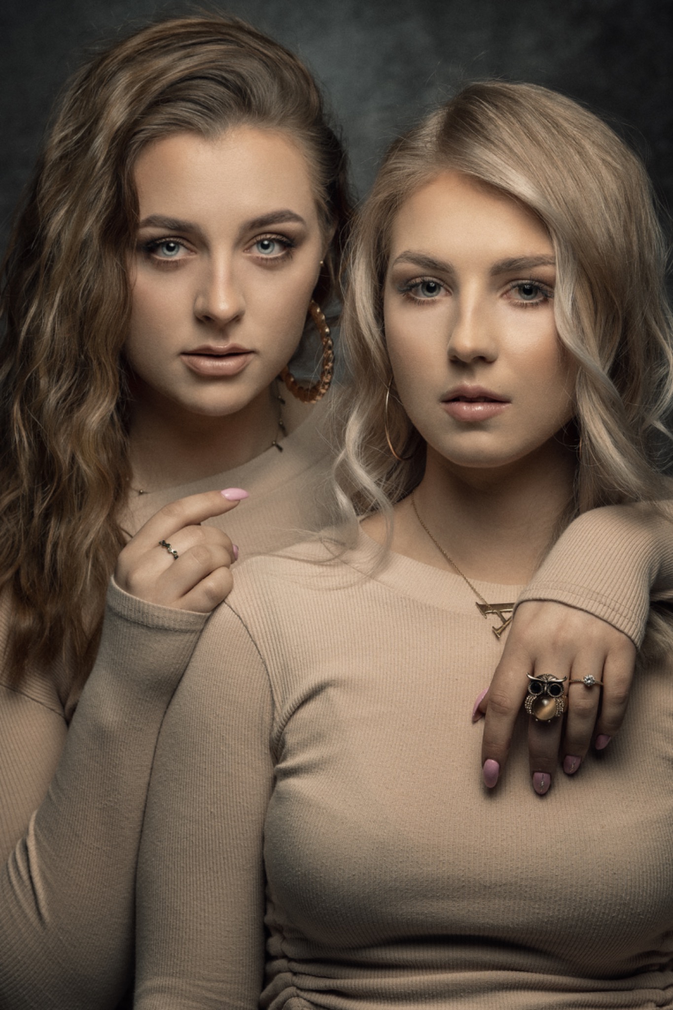 Portret dwóch młodych kobiet, blondynki i brunetki, w dopasowanych beżowych swetrach, pozujących na ciemnym tle, z wyraźnym akcentem na biżuterię i makijaż.