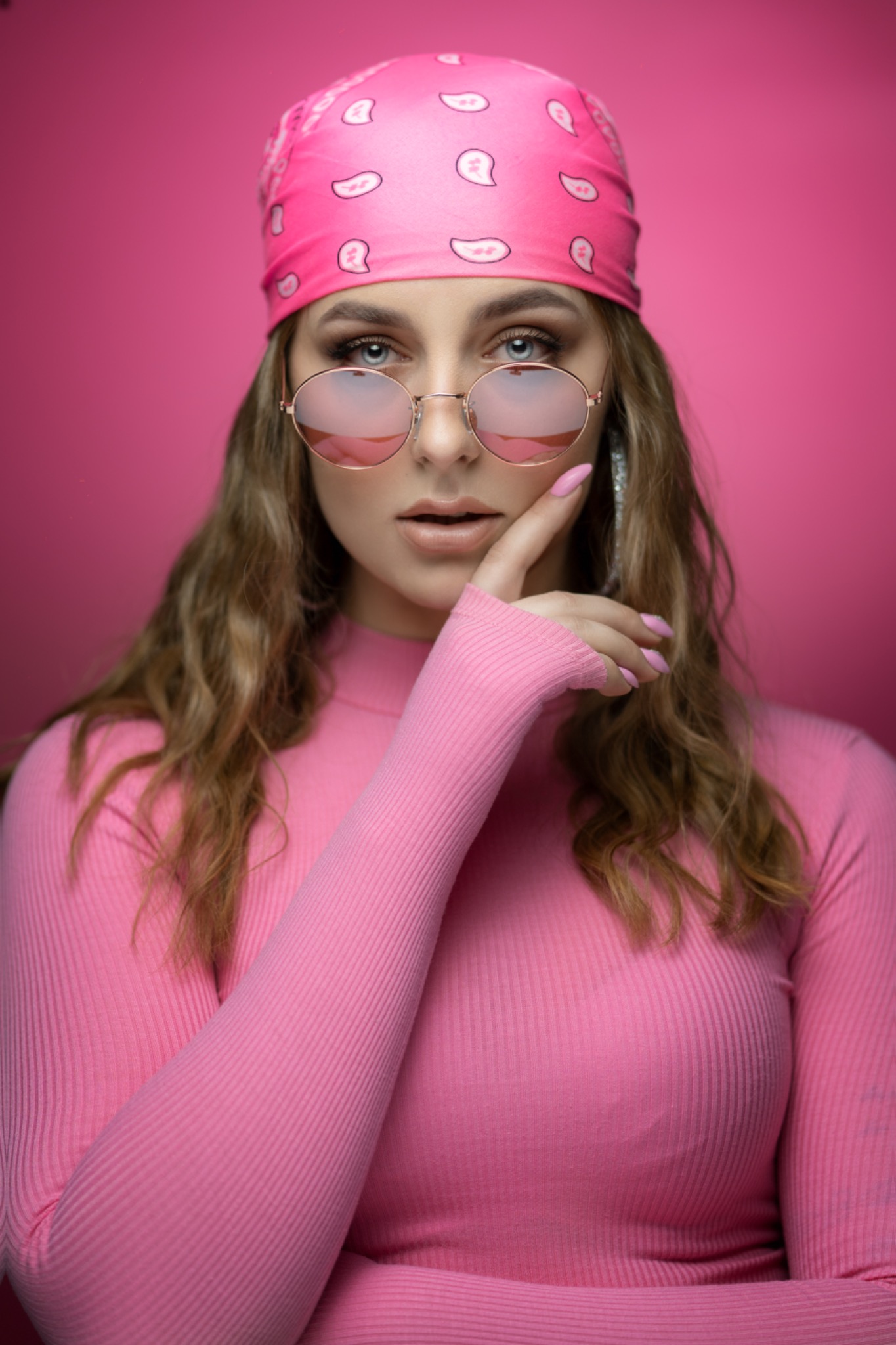Portret kobiety w różowym stroju i okularach, z dłonią przy twarzy na tle różowego tła.