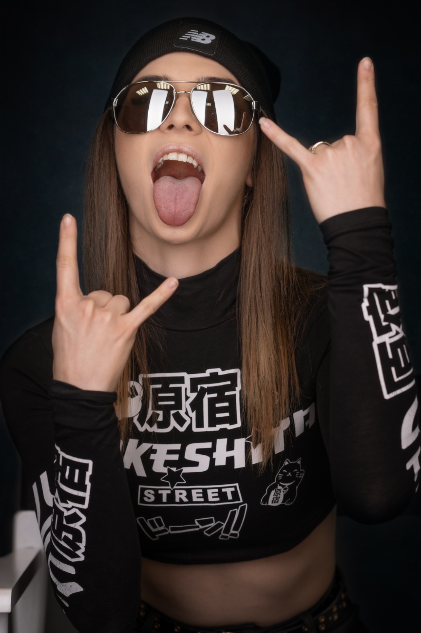 Portret młodej kobiety w czapce New Balance i okularach przeciwsłonecznych, wystawiającej język i pokazującej rockowy gest dłońmi, ubrana w czarny crop top z japońskimi napisami i rysunkiem kota.