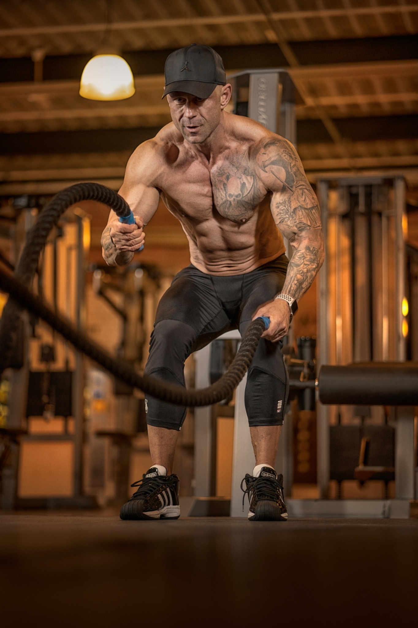 Mężczyzna z tatuażami, w czapce i sportowym stroju, intensywnie ćwiczy z linami w siłowni, eksponując muskulaturę.