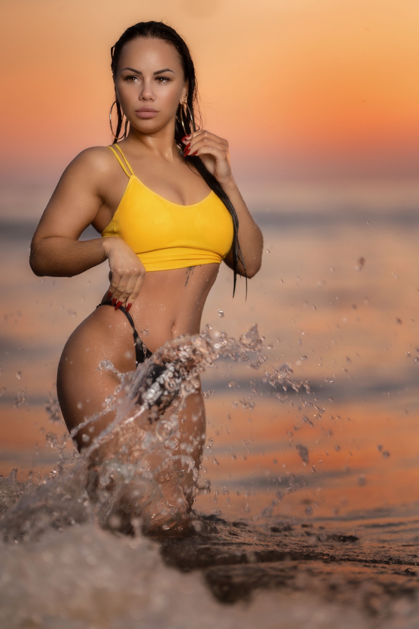 Kobieta w żółtym topie i czarnym bikini pozuje w wodzie podczas zachodu słońca, z kroplami wody w powietrzu.