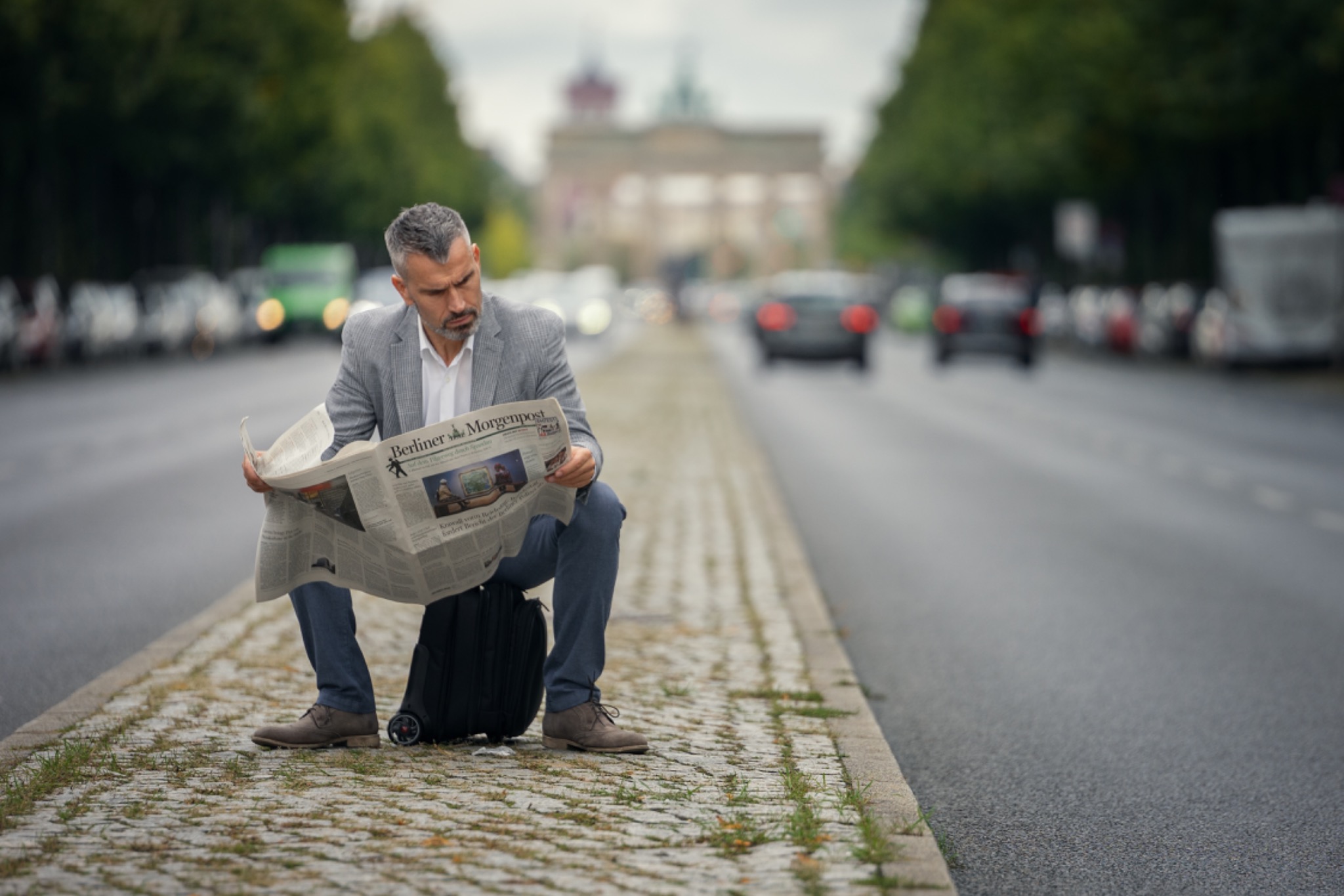Mężczyzna w Słupsku siedzi na walizce przy ulicy, czytając gazetę Berliner Morgenpost na tle Bramy Brandenburskiej.