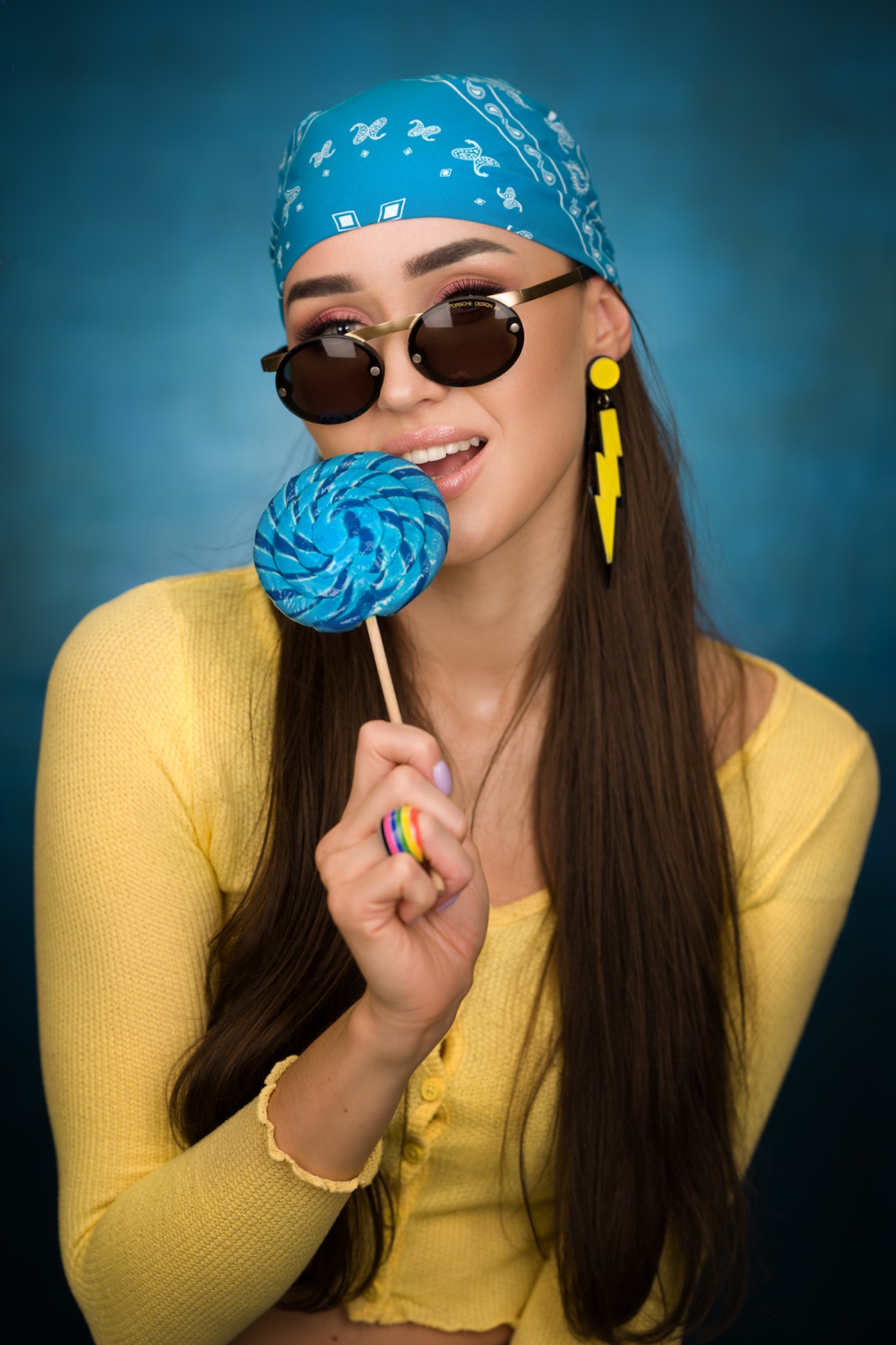 Portret kobiety w okularach przeciwsłonecznych i bandanie na głowie, trzymającej niebieskiego lizaka, z tęczowym pierścionkiem na palcu, na rozmytym niebieskim tle.