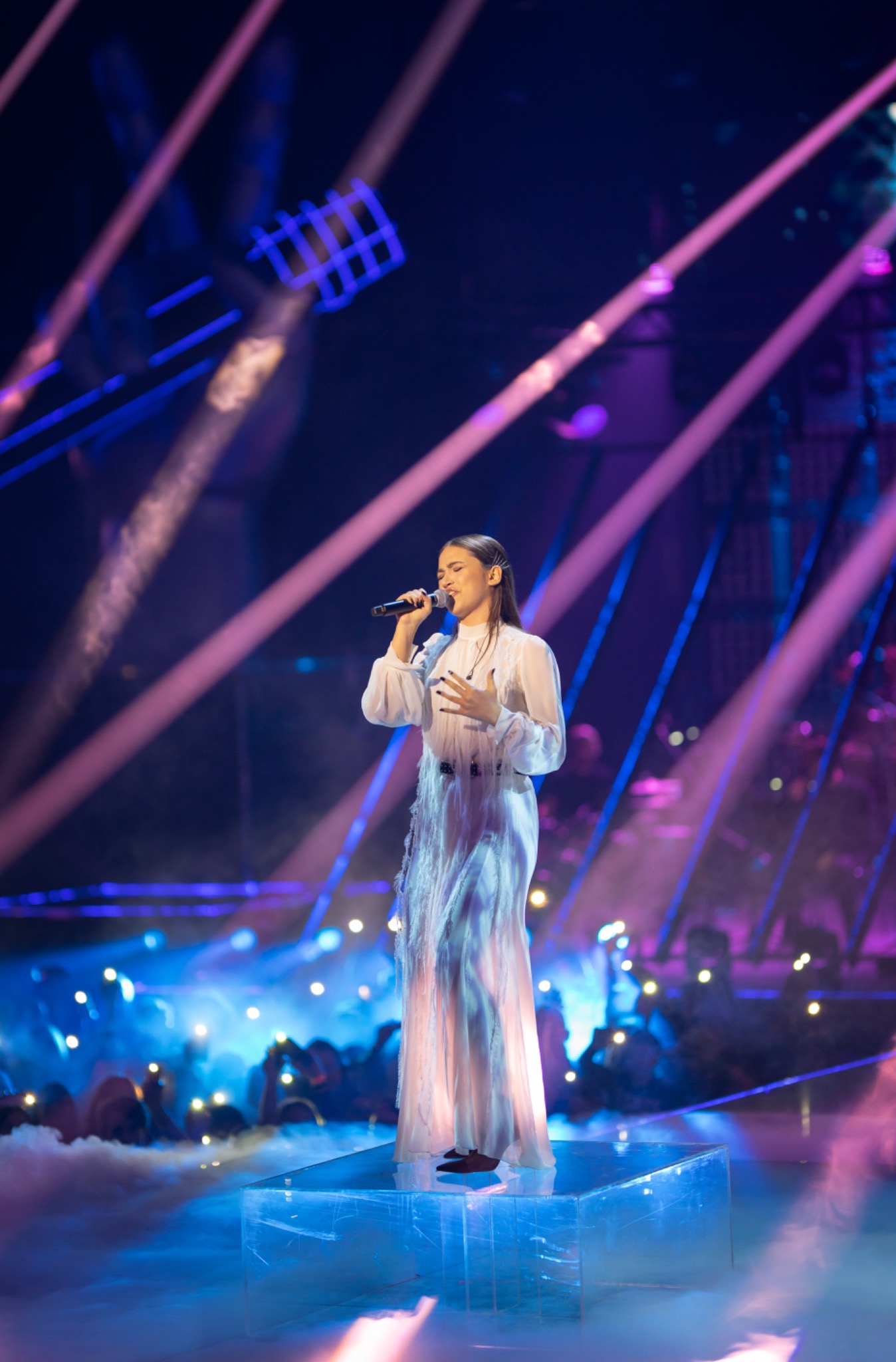 Młoda kobieta w białej sukni śpiewa do mikrofonu na podświetlonej scenie z publicznością w tle.