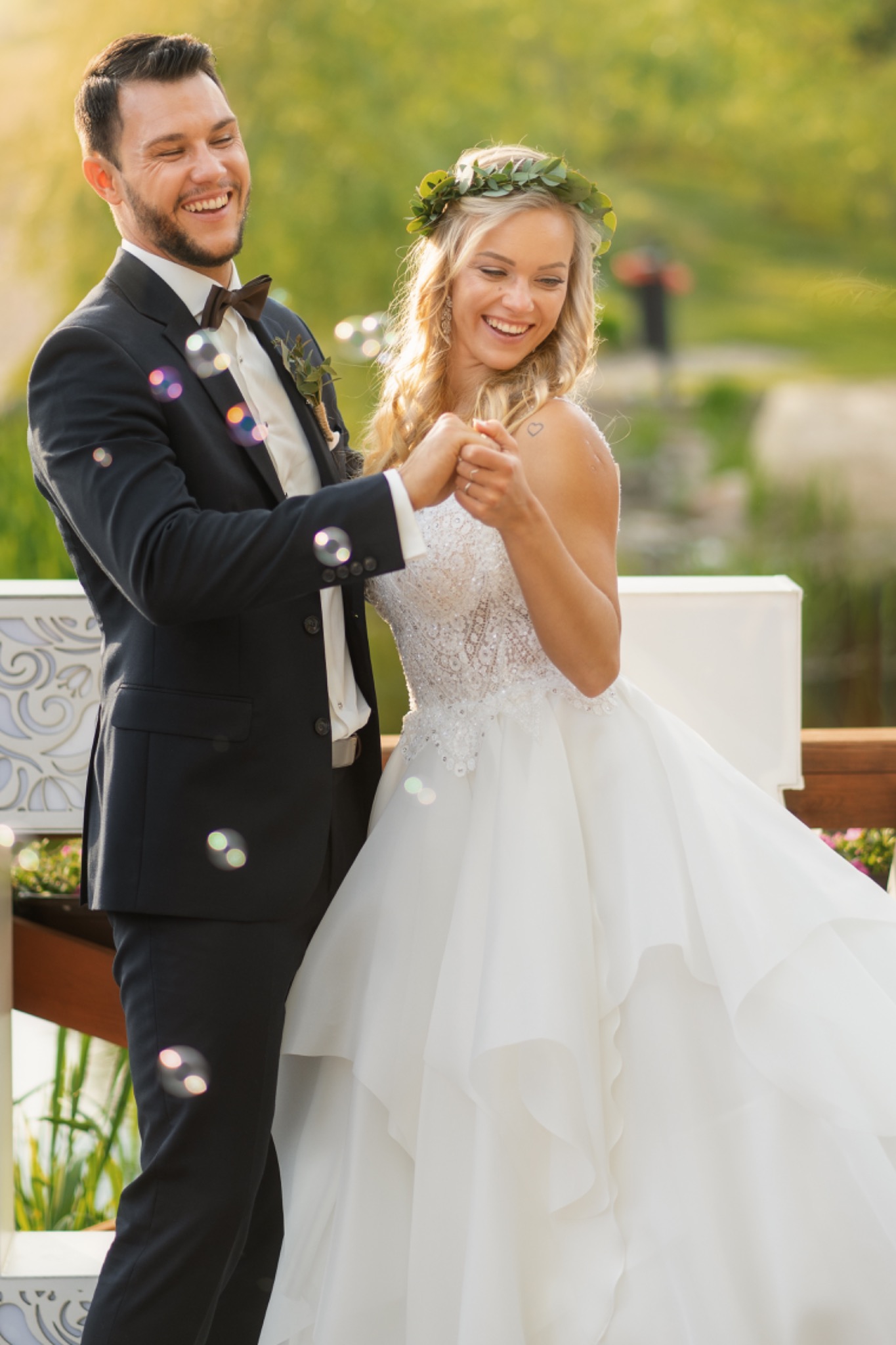 Szczęśliwa para młoda trzymająca się za ręce, otoczona bańkami mydlanymi; panna młoda w koronie z zielonych liści, pan młody w ciemnym garniturze z muszką.