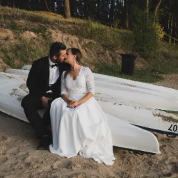 Para młoda w czułym pocałunku, siedząca na stosie kajaków na piaszczystej plaży, w tle las i żółte barierki schodów.