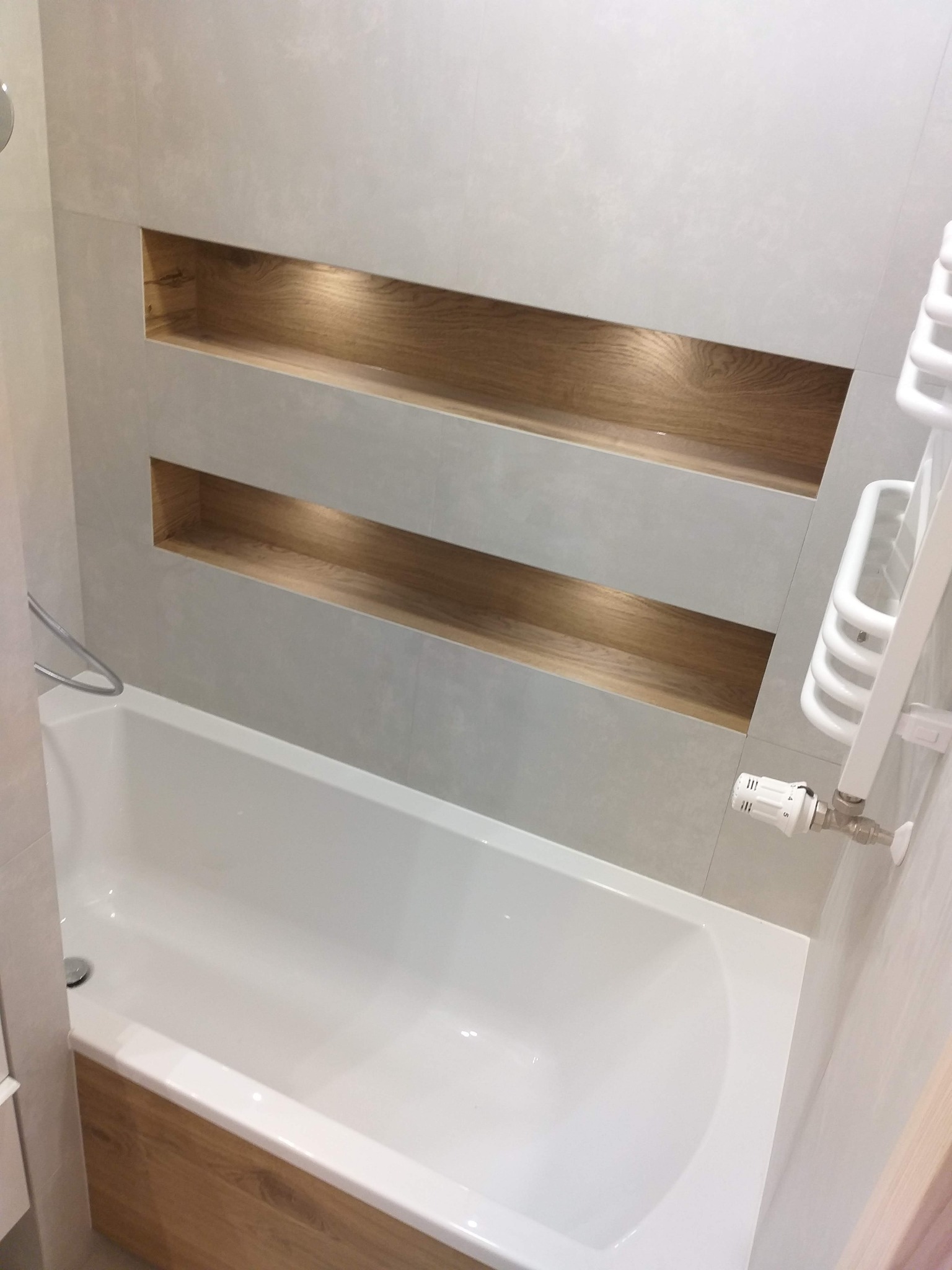 Nowoczesna łazienka z białą wanną obudowaną drewnem, wnęki ścienne z drewnianym wykończeniem i oświetleniem LED, grzejnik drabinkowy.