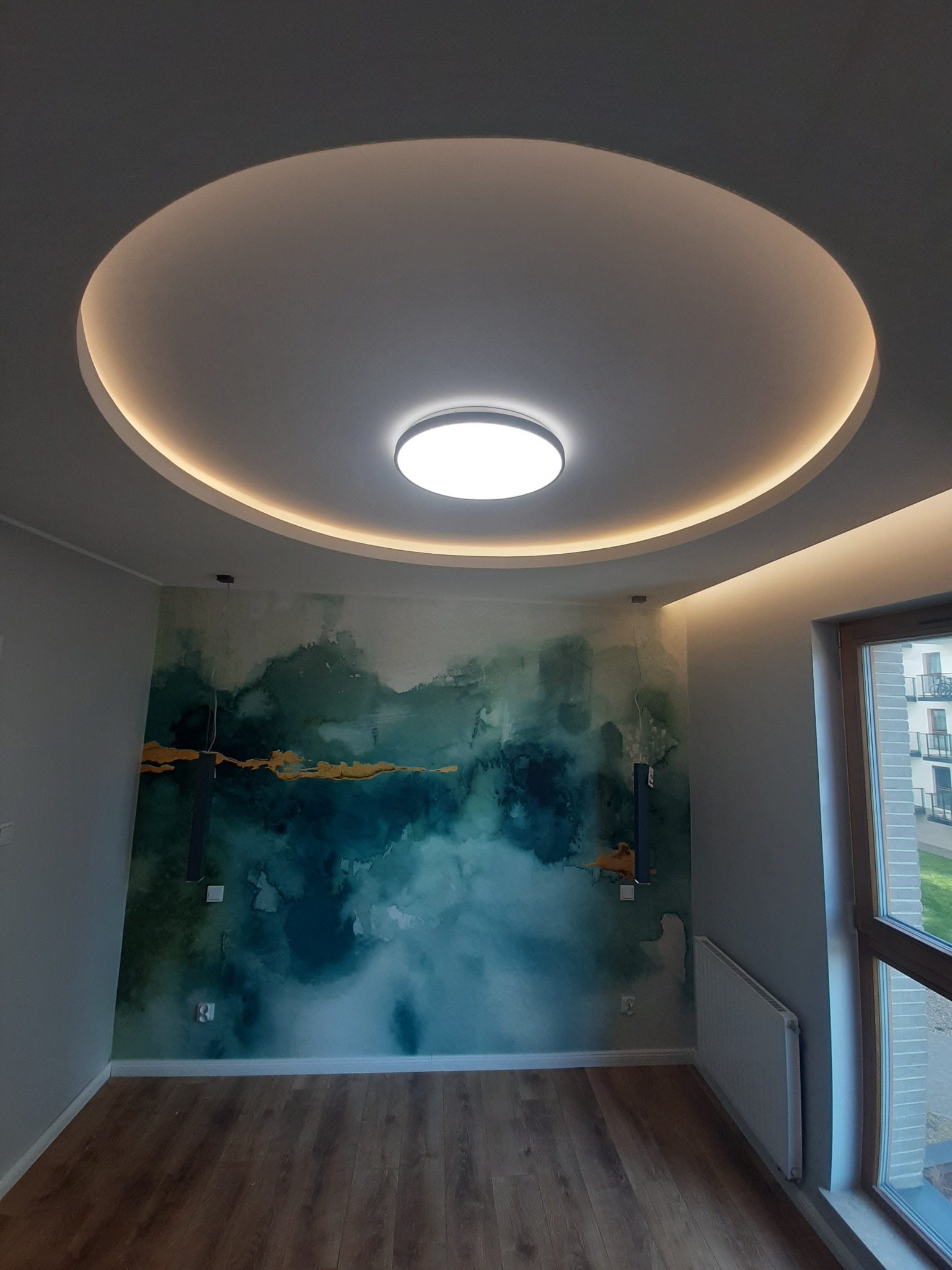 Nowoczesne wnętrze pokoju z dekoracyjną tapetą w odcieniach zieleni i złota, okrągłym sufitem podwieszanym z oświetleniem LED oraz panelami podłogowymi.