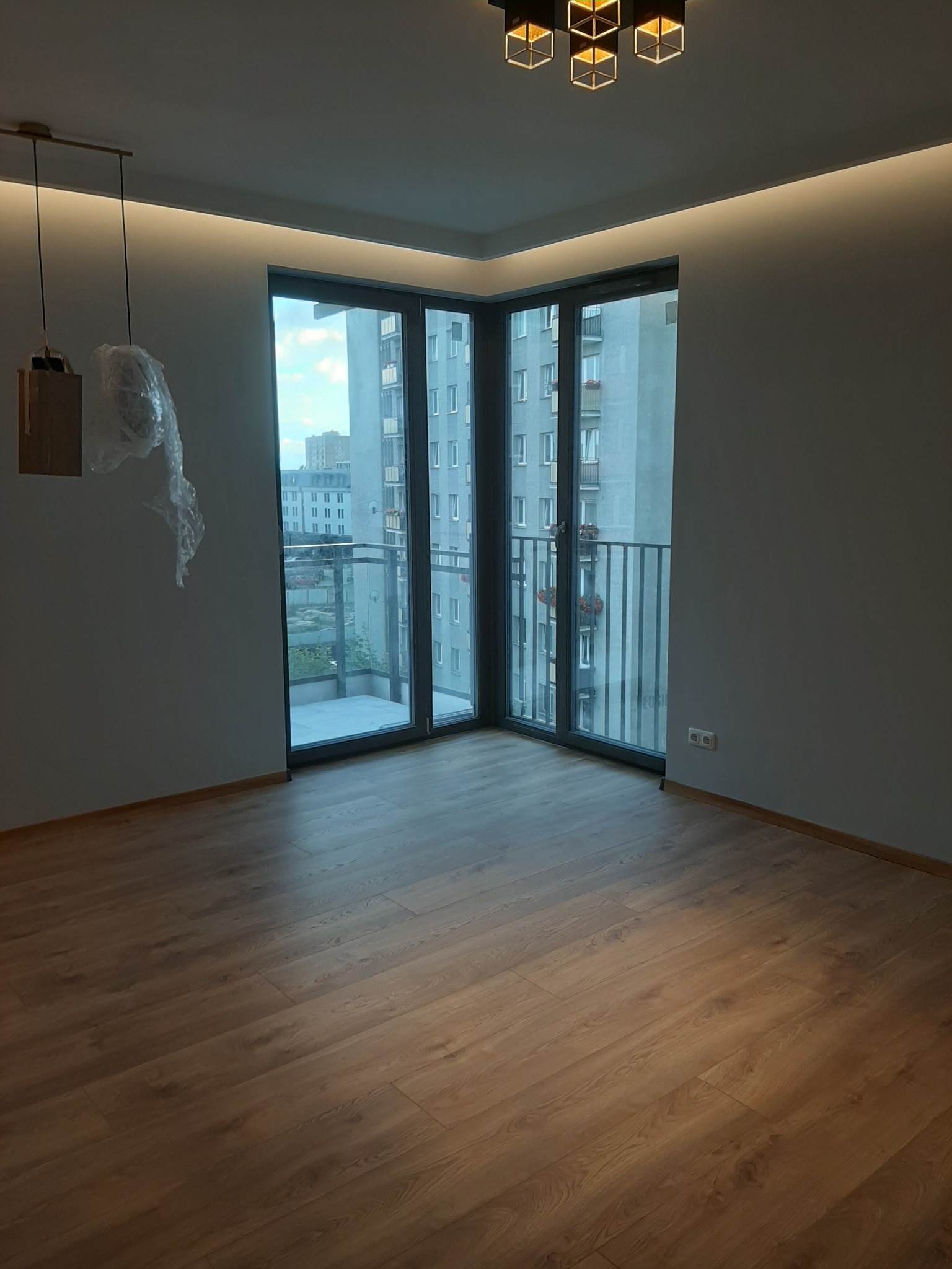 Nowoczesne wnętrze mieszkania po remoncie: panele podłogowe, okna balkonowe z widokiem na miasto, podwieszany sufit z oświetleniem LED, lampa wisząca zabezpieczona folią.