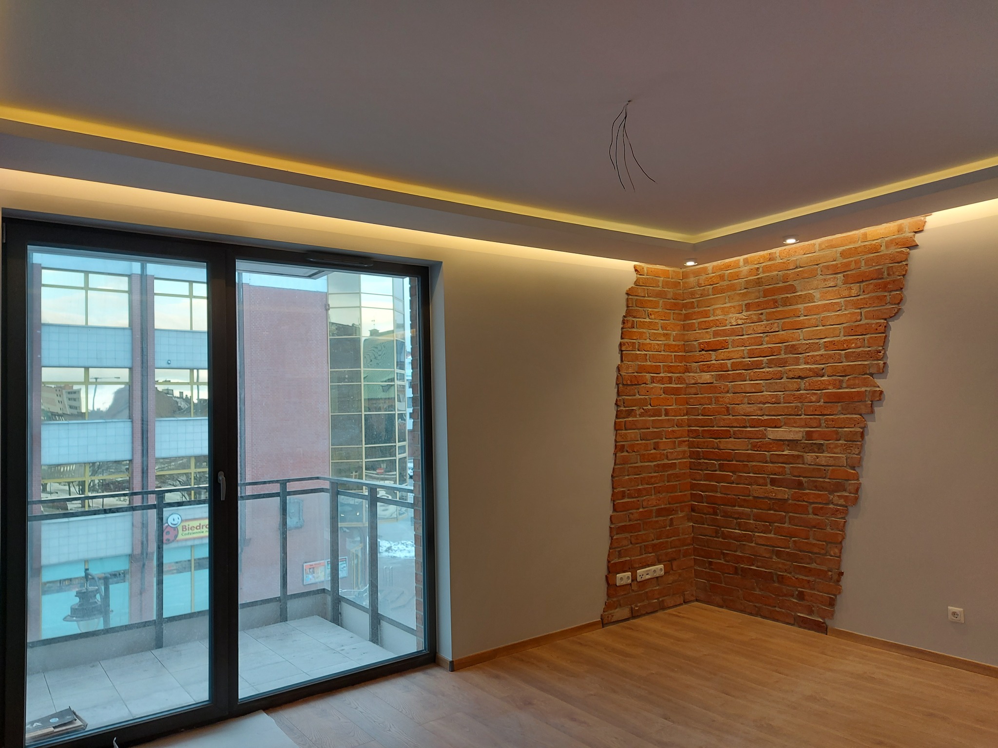 Pokój po remoncie z dekoracyjną ścianą z cegły, oświetleniem LED podsufitowym i panelami podłogowymi, widok z okna na budynek z balkonami.