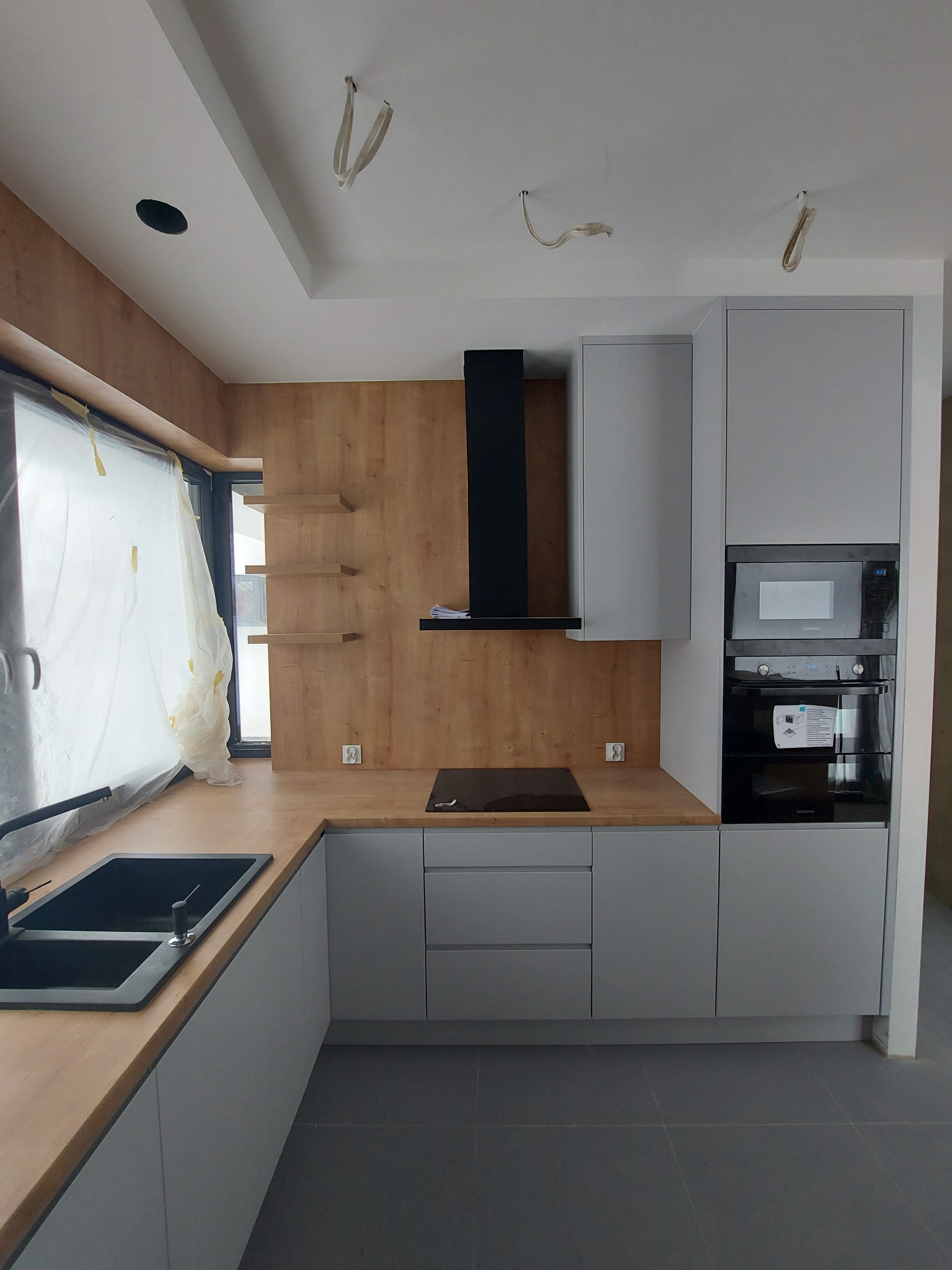 Nowoczesna kuchnia w szarościach i drewnie z czarnym zlewem i okapem, okno zabezpieczone folią malarską, widoczne niedokończone wykończenia sufitu.