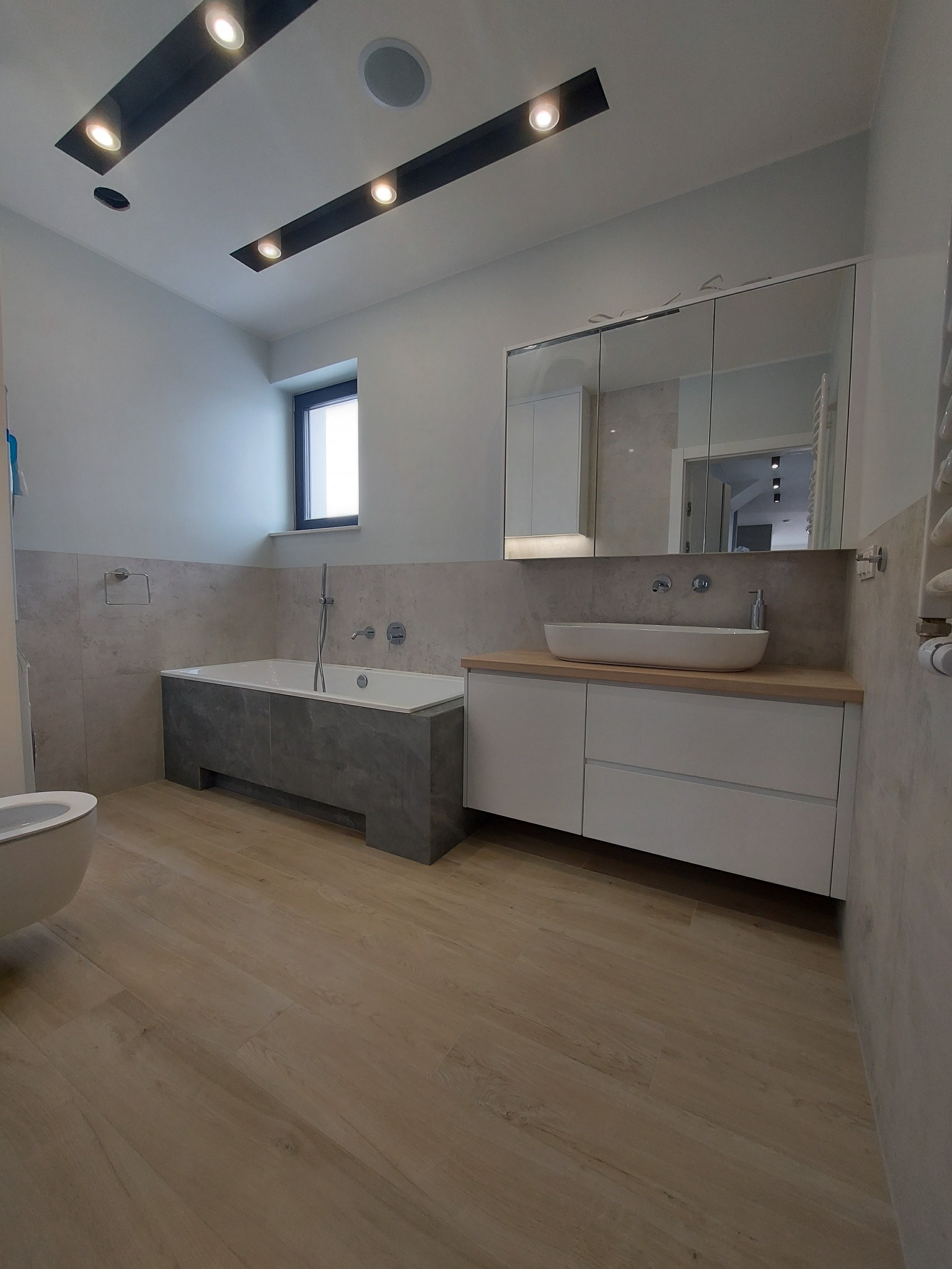 Nowoczesna łazienka z wanną obudowaną szarym kamieniem, białą umywalką nablatową na szafce z szufladami, lustrami w zabudowie i drewnopodobną podłogą.