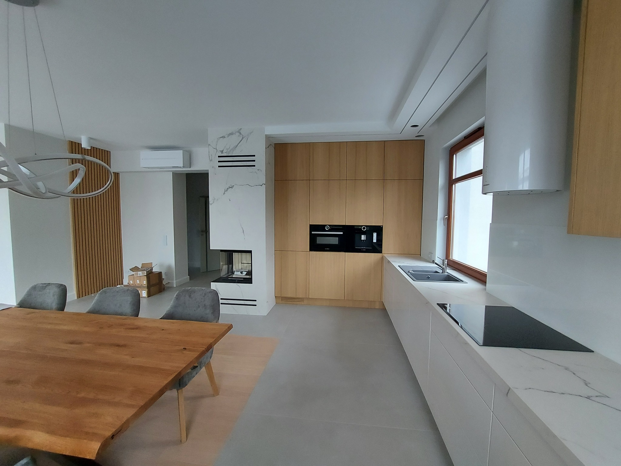 Nowoczesna kuchnia z drewnianymi szafkami, białym blatem z marmurowym wzorem, okapem cylindrycznym i drewnianym stołem jadalnym z szarymi krzesłami w tle