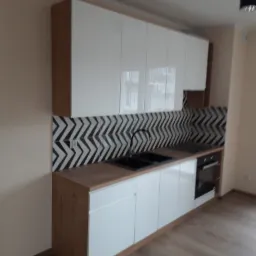 Nowoczesna kuchnia z białymi szafkami, drewnianym blatem i czarno-białymi płytkami w zygzaki nad blatem, widok na podłogę z paneli.