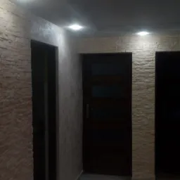 Korytarz po remoncie z imitacją kamienia na ścianach, gładzią na fragmencie ściany i szarą podłogą z płytek. Widoczne dwie ciemne, drewniane futryny drzwi i wbudowane oświetlenie sufitowe.