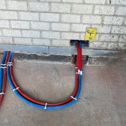 MOJO Damian Mojecki - Instalacja grzewcza podłogowa: czerwono-niebieskie rury przymocowane do podłoża, wchodzące w otwór w ścianie z cegły, obok żółta tabliczka informacyjna.