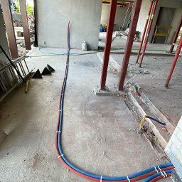 MOJO Damian Mojecki - Instalacja rur grzewczych (czerwona i niebieska) na surowym betonowym podłożu w trakcie budowy. Widoczne podpory stropu i elementy konstrukcyjne.