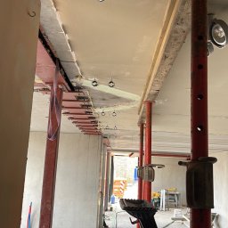 MOJO Damian Mojecki - Instalacja rur w nowym budynku, widoczne czerwone i niebieskie rury pod sufitem, zamocowane za pomocą uchwytów. Czerwone podpory budowlane i narzędzia w tle.