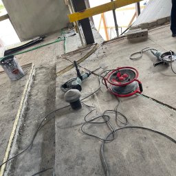 MOJO Damian Mojecki - Przygotowanie podłoża pod instalacje: bruzdy w betonie, narzędzia (wiertarka, szlifierka) i przedłużacz na podłodze. Widok na zewnątrz przez niezabezpieczony otwór.