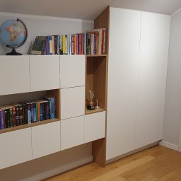 INSIDE Anna Weyna - Biała zabudowa meblowa z elementami drewnopodobnymi, obejmująca szafę i regał z półkami na książki, globus oraz dekoracje, uzupełniona o białe drzwi z poziomymi szarymi paskami.