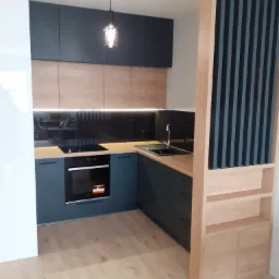 Nowoczesna kuchnia z ciemnymi, gładkimi szafkami górnymi i drewnopodobnymi frontami dolnymi, blatem w odcieniu drewna oraz czarnym panelem szklanym nad blatem, oświetlona wiszącą lampą i listwą LED...