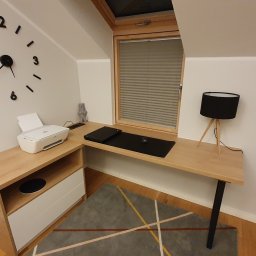 INSIDE Anna Weyna - Biurko narożne z jasnego drewna pod oknem dachowym, z drukarką, lampką i dużym zegarem ściennym.