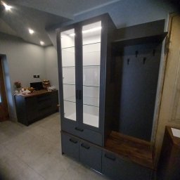 INSIDE Anna Weyna - Szara zabudowa meblowa z przeszkloną witryną i siedziskiem z wieszakami w przedpokoju, widok na komodę w tle.