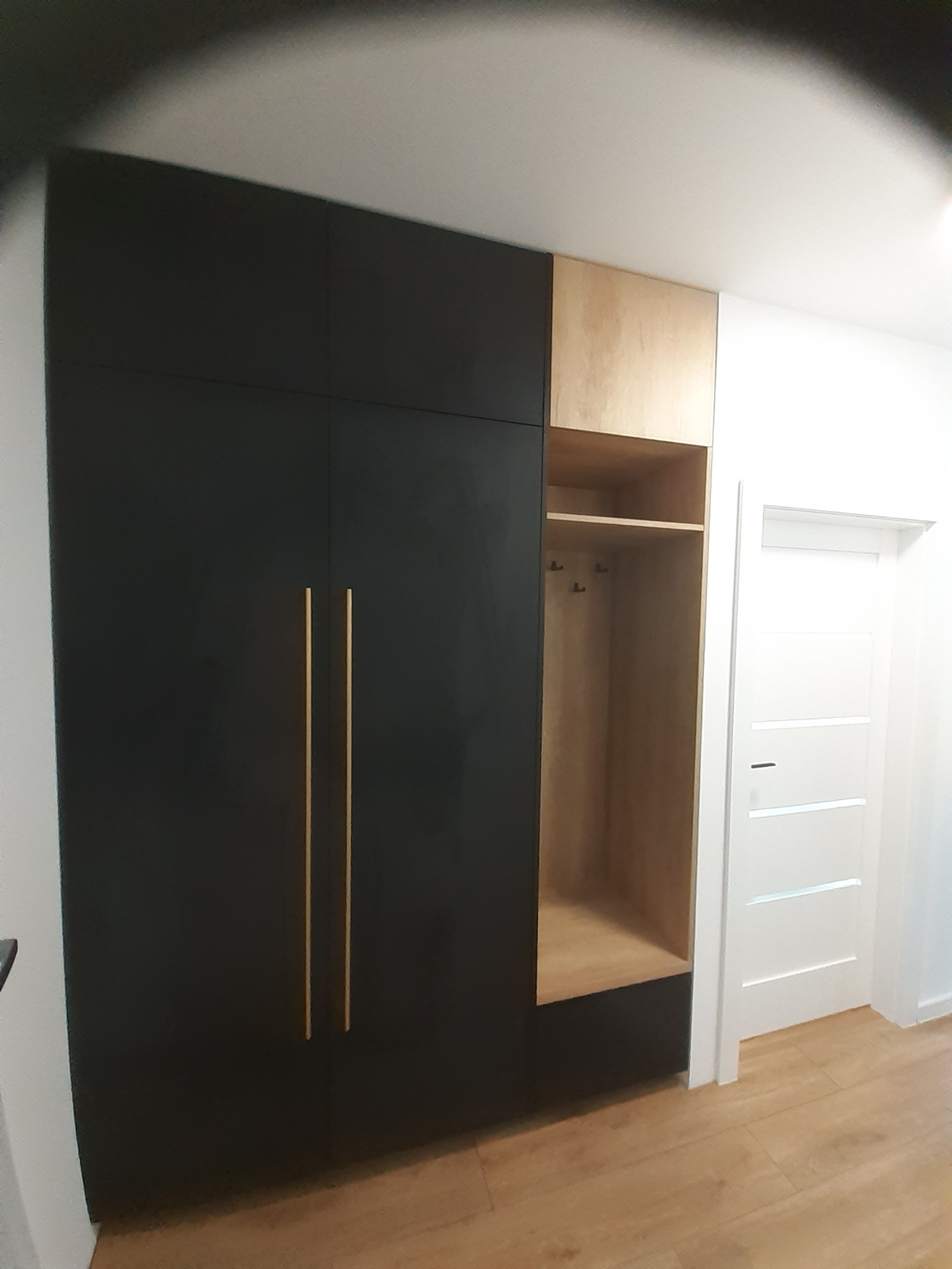 Czarno-drewniana szafa wnękowa z otwartą półką i złotymi uchwytami, widok z boku na tle białej ściany i drzwi.