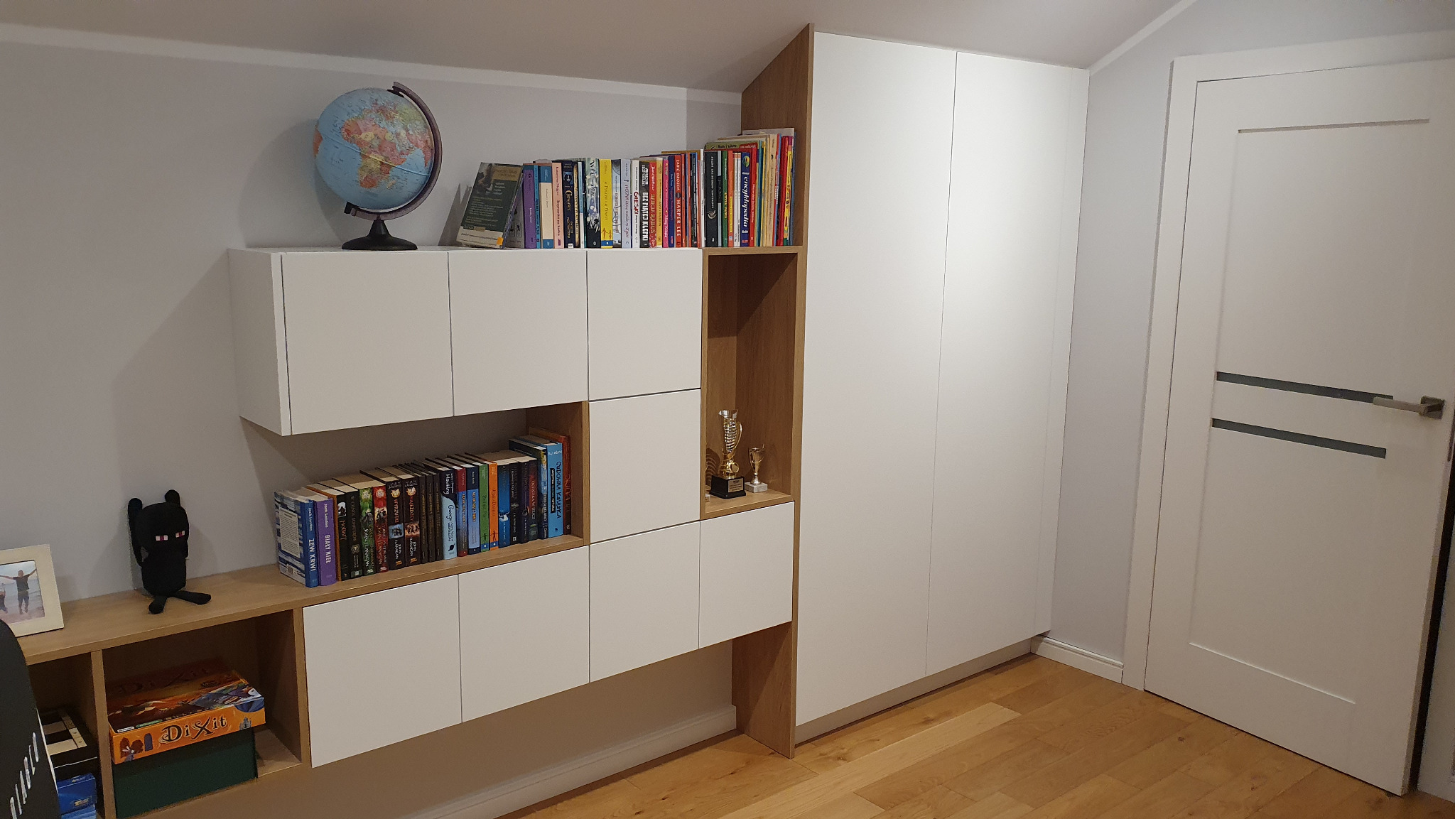 Biała zabudowa meblowa z elementami drewnopodobnymi, obejmująca szafę i regał z półkami na książki, globus oraz dekoracje, uzupełniona o białe drzwi z poziomymi szarymi paskami.