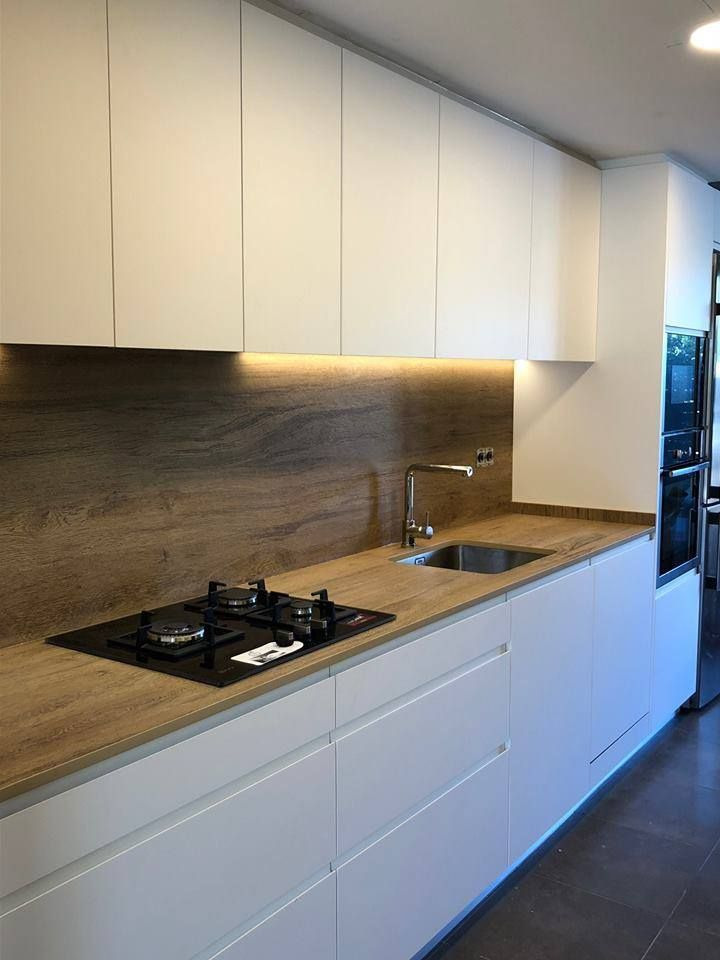 Nowoczesna kuchnia z białymi, gładkimi frontami szafek i blatem imitującym drewno, oświetlonym od spodu, z czarną płytą gazową i stalowym zlewem.