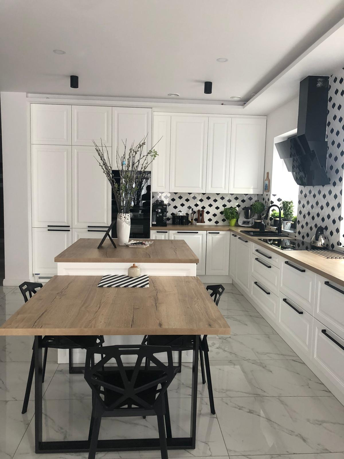 Nowoczesna biała kuchnia z drewnianym blatem i wyspą, czarne akcenty w postaci armatury, okapu i krzeseł, marmurowa podłoga, dekoracyjne backsplash w romby.