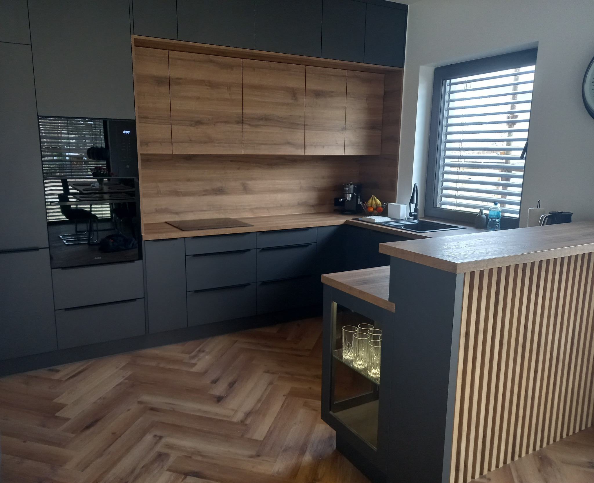 Nowoczesna kuchnia z szarymi szafkami dolnymi i drewnianymi górnymi, blatem z imitacją drewna i barkiem z lamelami, na podłodze parkiet w jodełkę.