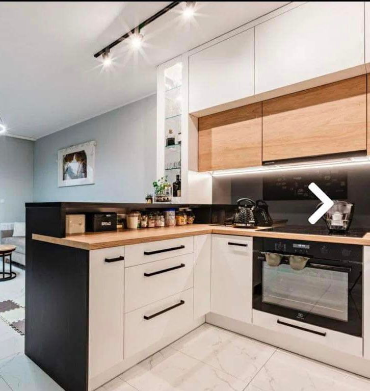 Nowoczesna kuchnia w odcieniach bieli, czerni i drewna z czarnymi, podłużnymi uchwytami, oświetleniem podszafkowym LED i marmurową podłogą.