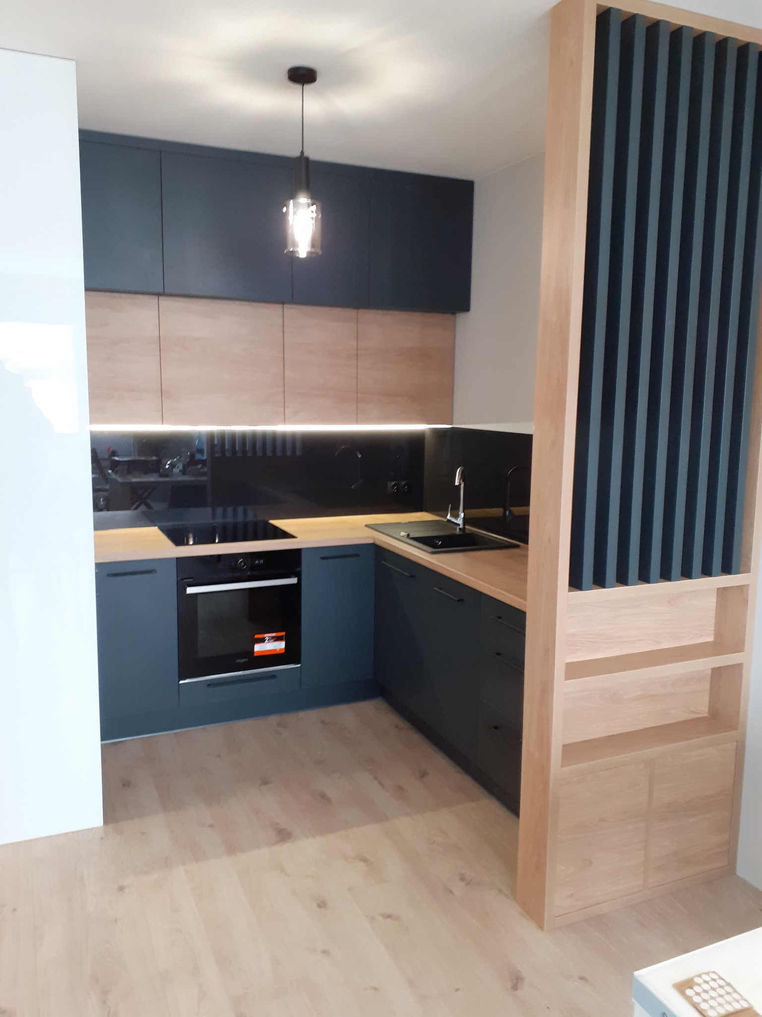 Nowoczesna kuchnia z ciemnymi, gładkimi szafkami górnymi i drewnopodobnymi frontami dolnymi, blatem w odcieniu drewna oraz czarnym panelem szklanym nad blatem, oświetlona wiszącą lampą i listwą LED...