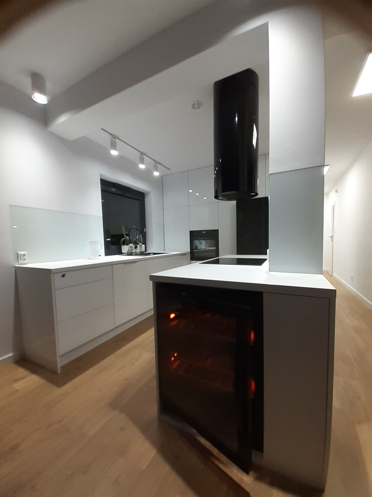 Minimalistyczna, biała kuchnia z wyspą i wbudowaną chłodziarką do wina, oświetlona reflektorami sufitowymi, z czarnym okapem cylindrycznym.