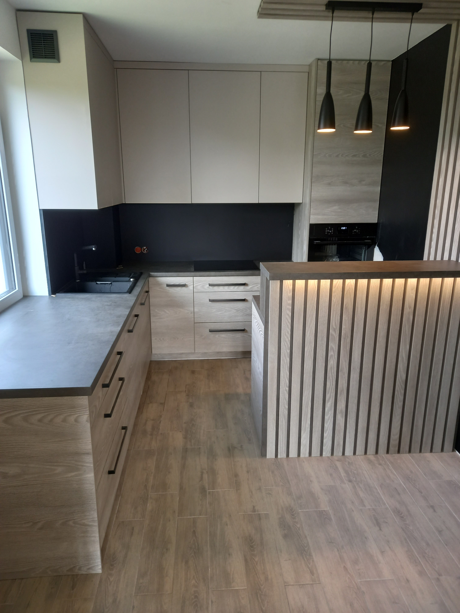 Nowoczesna kuchnia z drewnianymi frontami, czarnym blatem i oświetleniem punktowym nad półwyspem z lamelami.