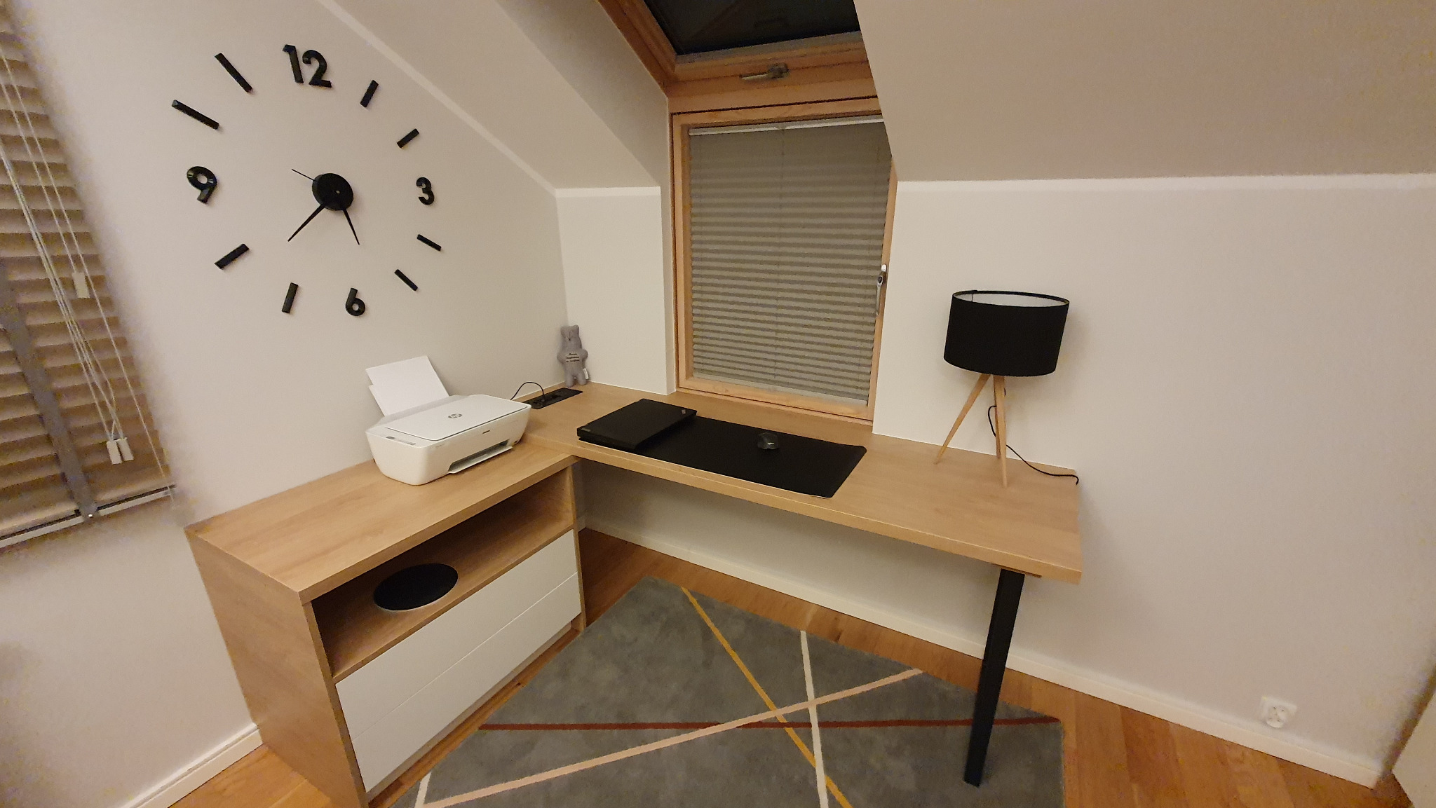 Biurko narożne z jasnego drewna pod oknem dachowym, z drukarką, lampką i dużym zegarem ściennym.