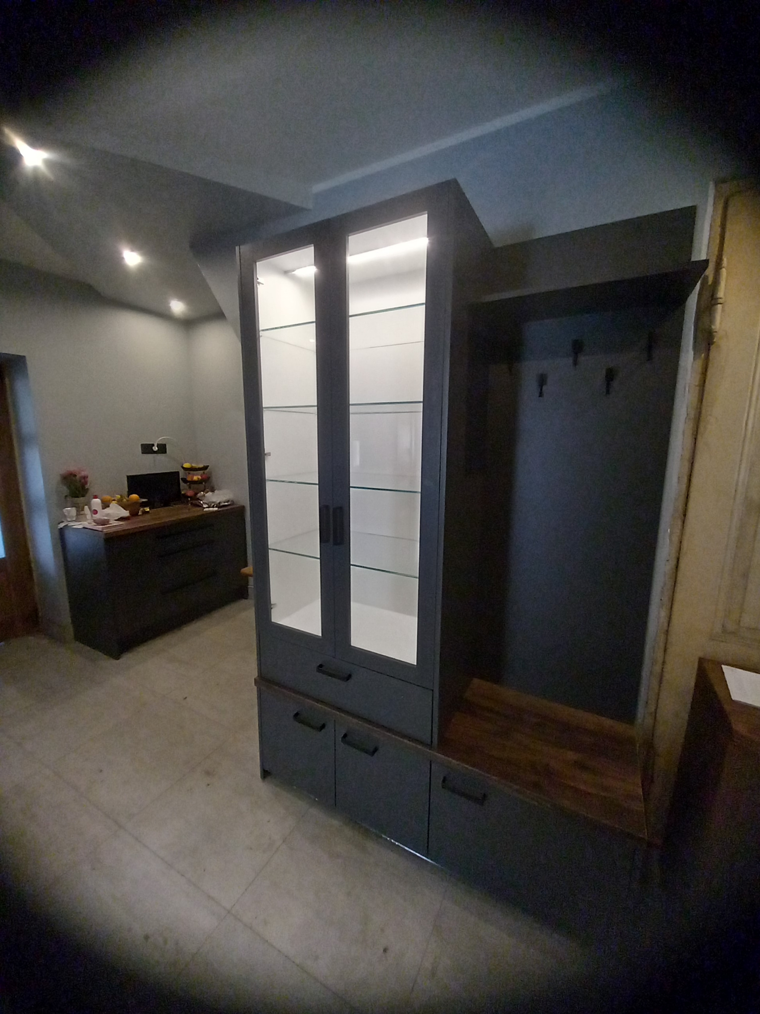 Szara zabudowa meblowa z przeszkloną witryną i siedziskiem z wieszakami w przedpokoju, widok na komodę w tle.