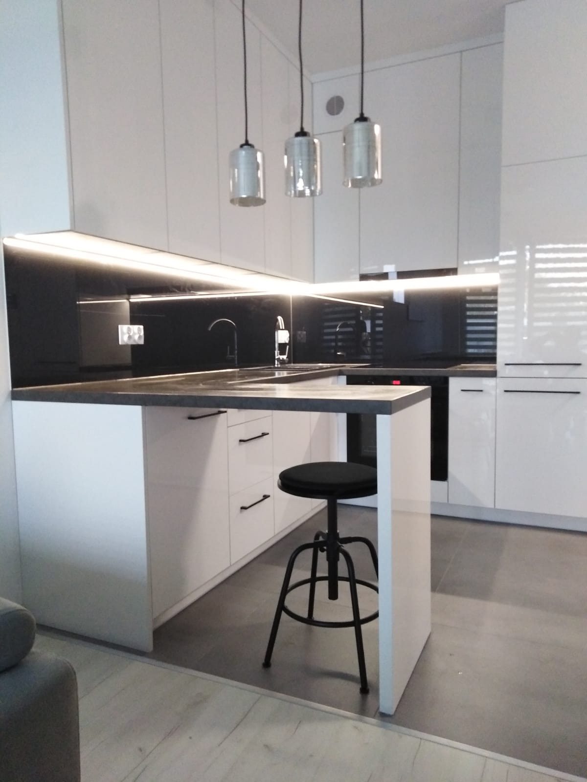Biała kuchnia z czarnym blatem, oświetleniem LED pod szafkami i trzema wiszącymi lampami nad wyspą kuchenną z czarnym stołkiem barowym na metalowej nodze.