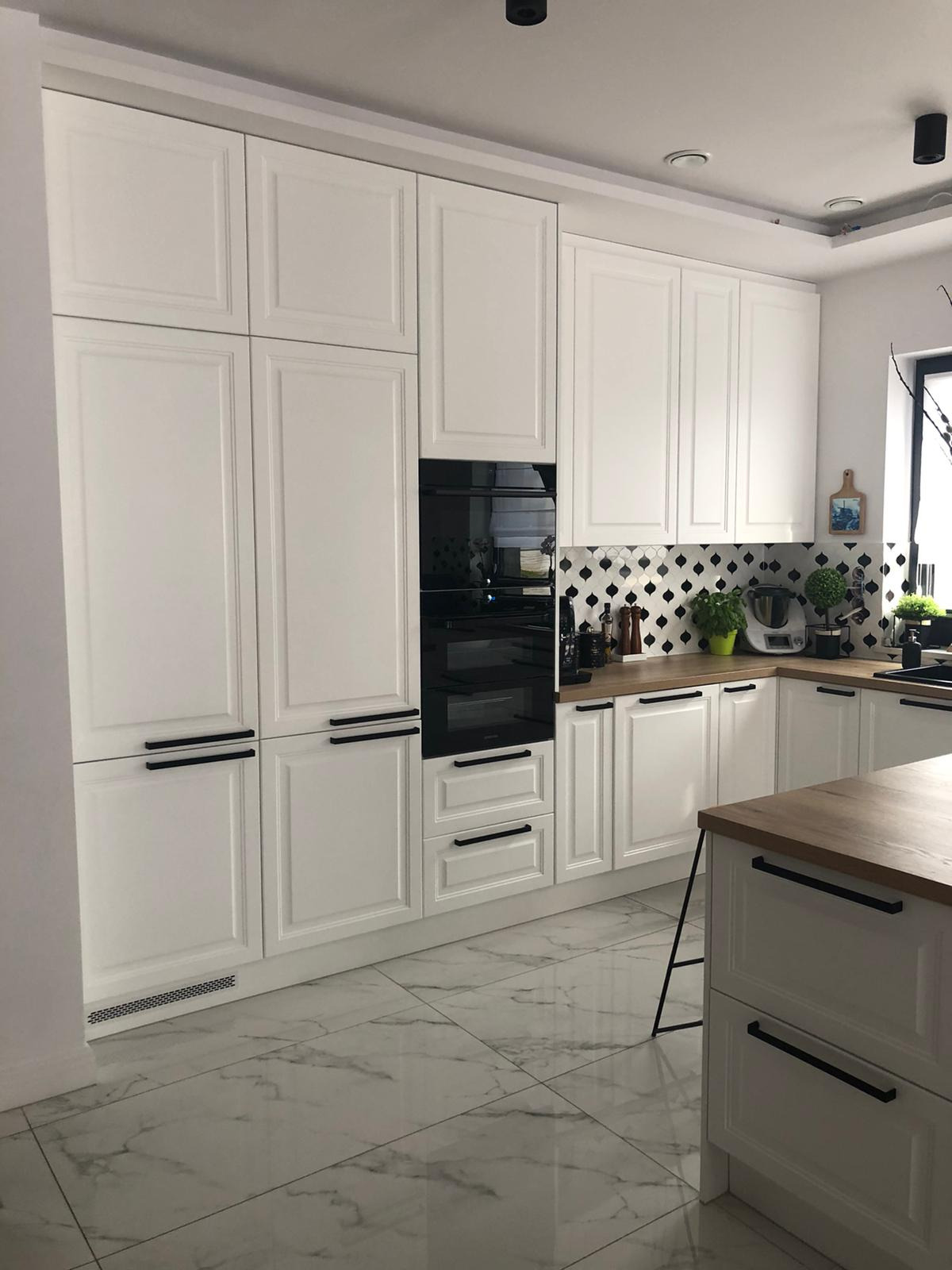 Biała kuchnia z frezowanymi frontami, czarnymi, długimi uchwytami, drewnianym blatem i marmurową podłogą, oświetlona światłem dziennym.