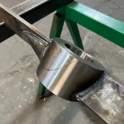 Detal spawania elementu metalowego z okrągłym otworem, widoczne spoiny i szlifowana powierzchnia, na tle warsztatowego stołu.