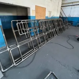 Stos metalowych konstrukcji rurowych, potencjalnie elementów balustrad, ułożonych w warsztacie z widocznym okablowaniem i surowymi ścianami.