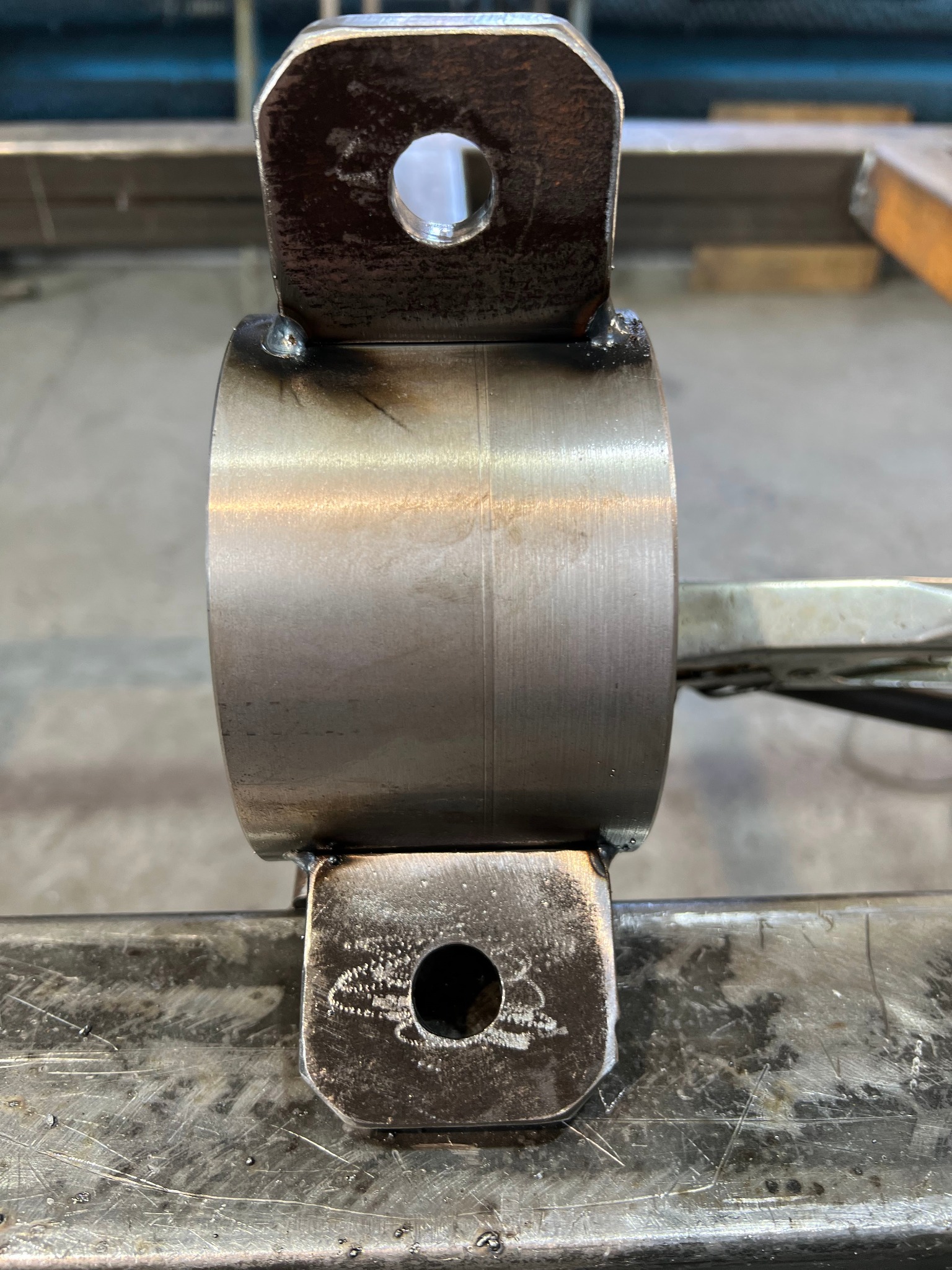 Metalowy element z przyspawanymi uchwytami z otworami, widoczne spawy i ślady obróbki na powierzchni metalu.