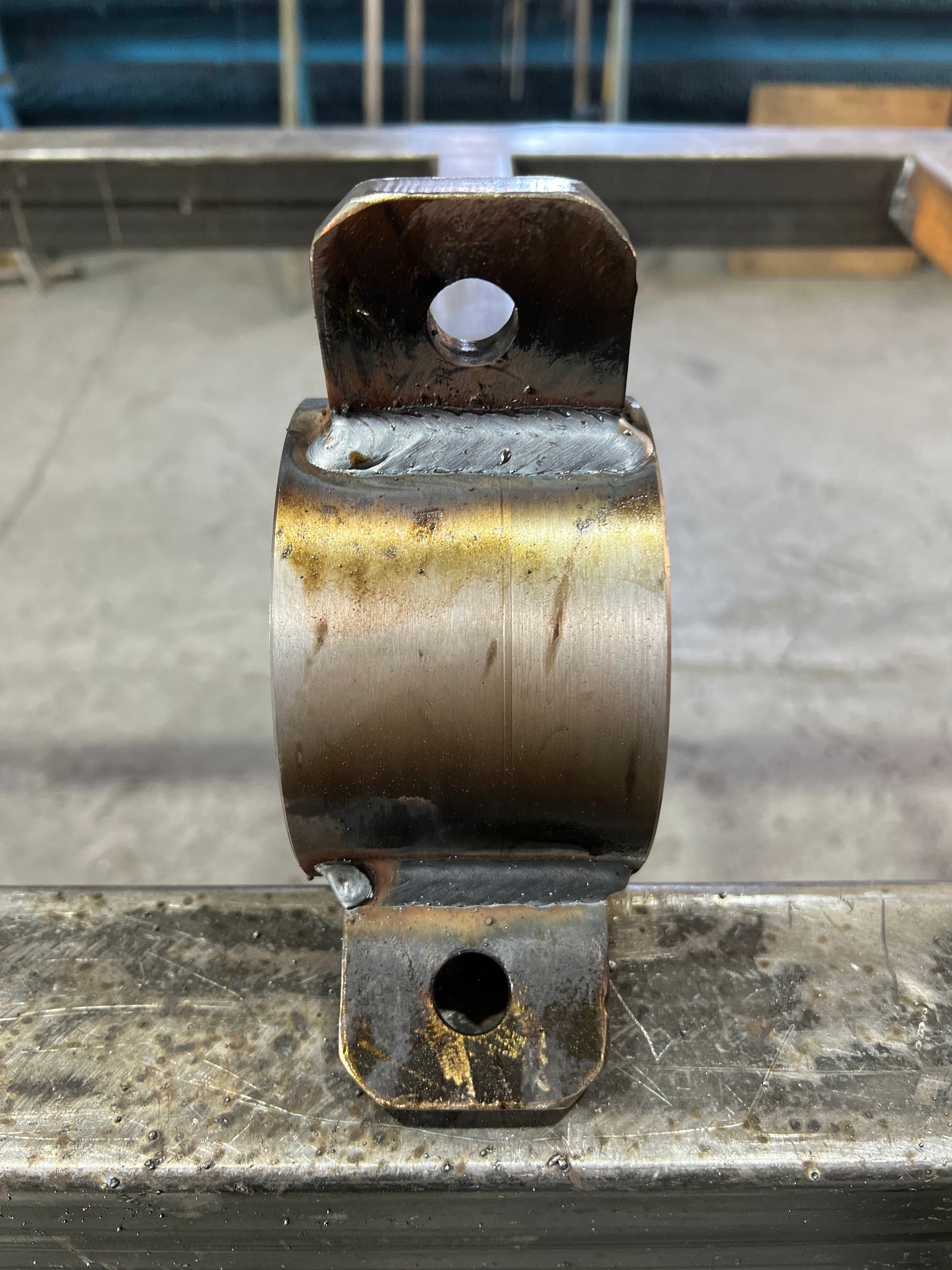 Metalowy uchwyt z przyspawanymi elementami mocującymi, widoczne spawy i otwory montażowe, umieszczony na metalowej konstrukcji.