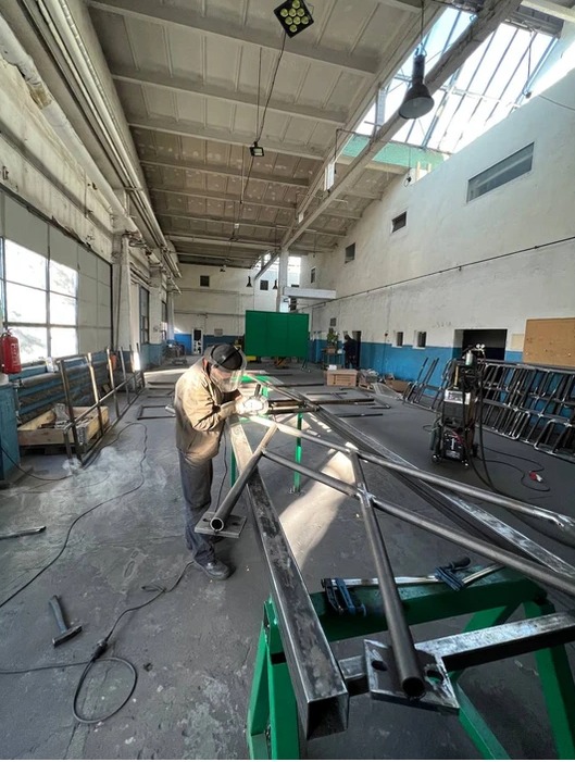 Pracownik w kombinezonie i masce spawalniczej łączy metalowe rury w konstrukcji przypominającej fragment balustrady w hali produkcyjnej z surowym betonowym stropem i świetlikami.