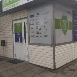 Zewnętrzny widok biura ubezpieczeń z szyldem 'Ubezpieczenia' i logo różnych towarzystw ubezpieczeniowych na oknach, w tym Compensa, Aviva, Ergo Hestia, PZU, Warta, HDI. Na szyldzie informacja...