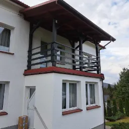 Balkon z czarną drewnianą balustradą i zadaszeniem, biała elewacja budynku z widocznymi oknami i kartonami przy drzwiach wejściowych.