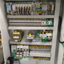 Wnętrze szafy sterowniczej z widocznymi elementami elektrycznymi, takimi jak przekaźniki, bezpieczniki, transformatory, listwy zaciskowe i okablowanie w kolorach czarnym, ż&oacute;łtym i zielonym...