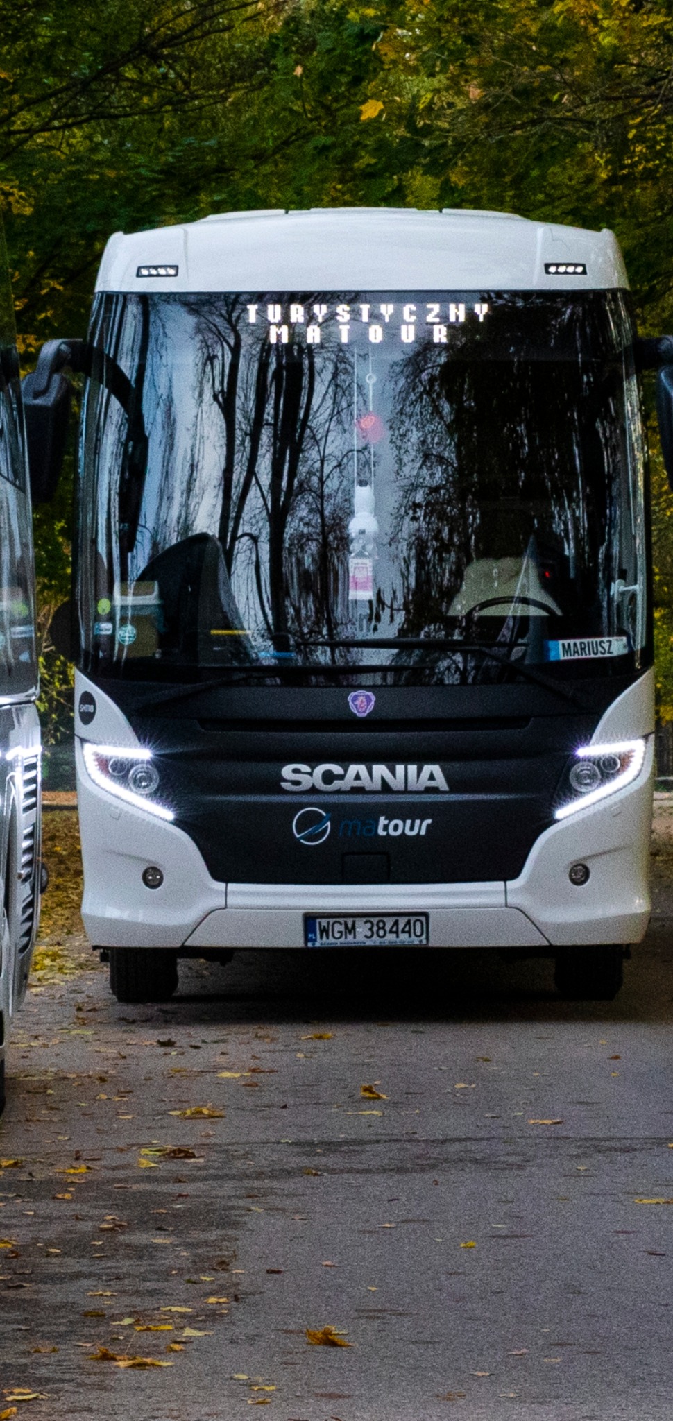 Biały autokar Scania 'Matour' z włączonymi światłami dziennymi, zaparkowany na parkingu z widocznymi opadłymi liśćmi, w tle drzewa