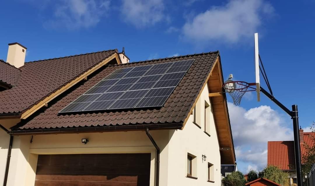 Panele słoneczne na brązowym dachu dwuspadowym domu jednorodzinnego z garażem, na tle błękitnego nieba z chmurami oraz kosz do koszykówki po prawej stronie.